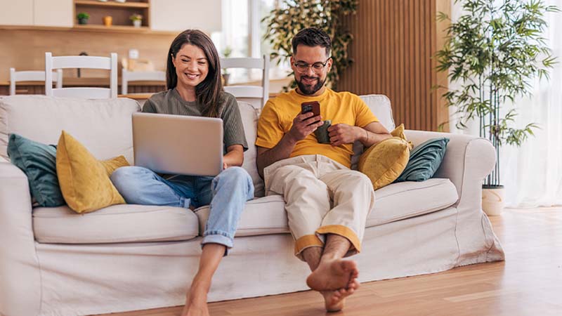 Duas pessoas sentadas num sofá claro numa sala acolhedora; uma usa um portátil e a outra utiliza um telemóvel, com almofadas e plantas ao redor.
